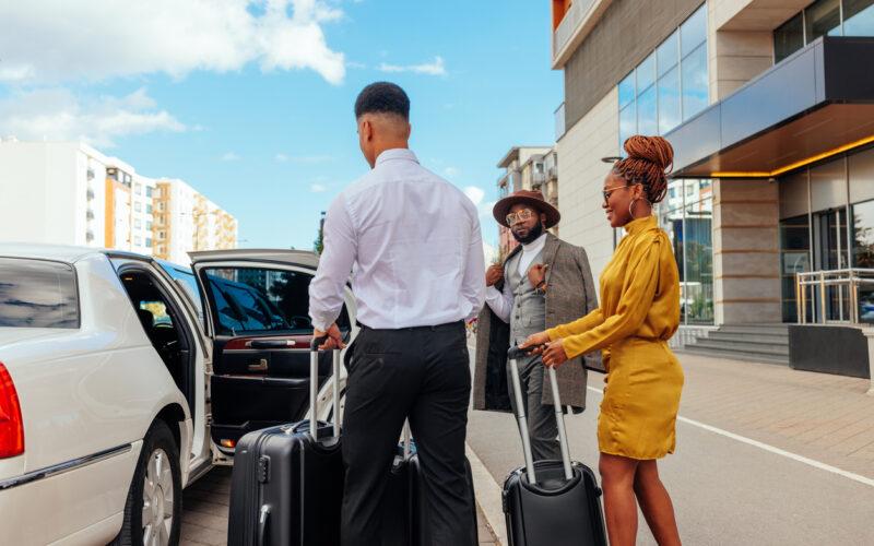 A limo driver is helping a wealthy couple with their luggage and into the car. They are well dresses and leaving a luxurious hotel.
